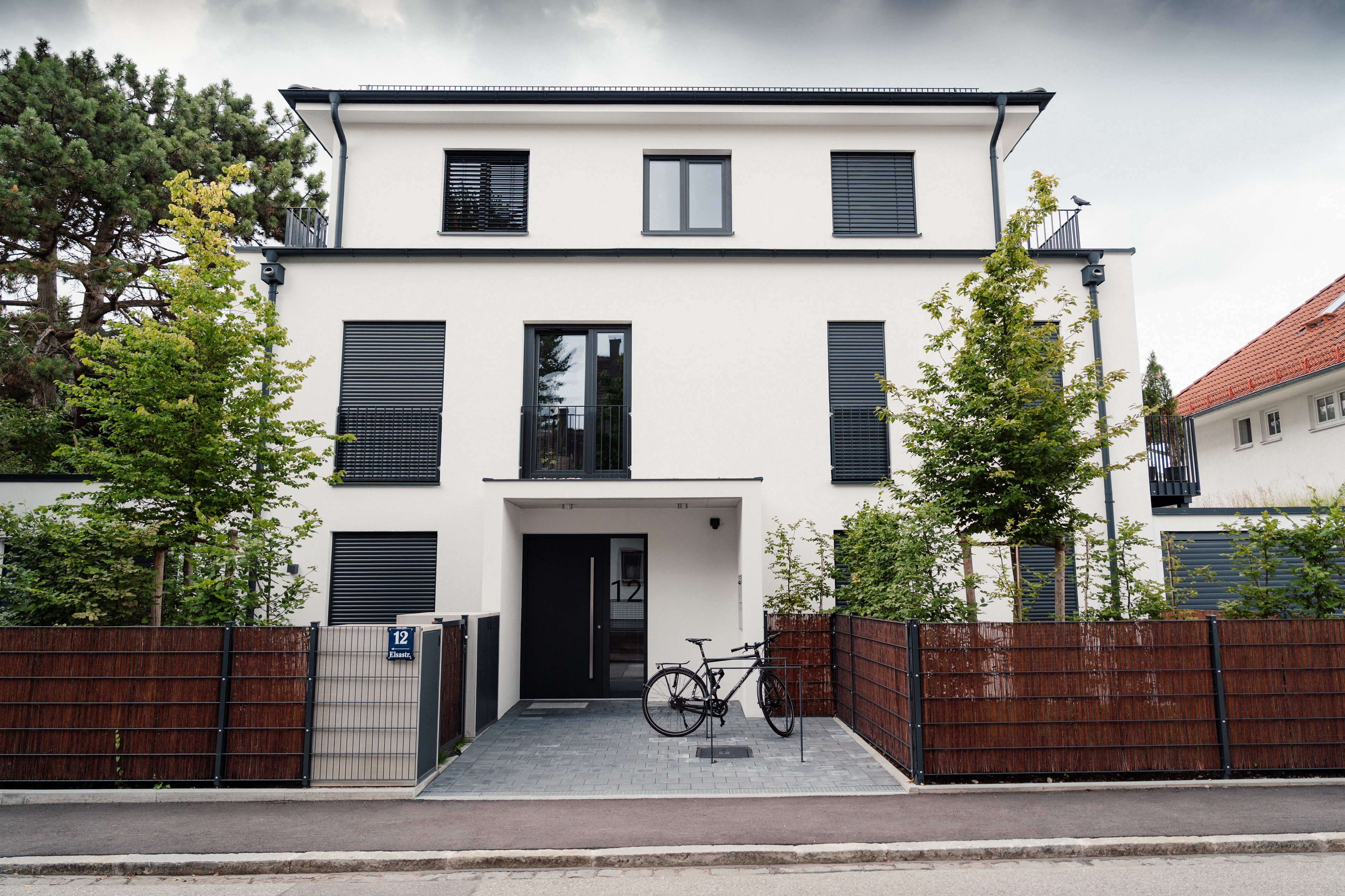 Elsastraße 12 – Frontansicht des Mehrfamilienhauses mit Eingangsvordach, Zaun, jungen Bäumen und Fahrrad am Eingang