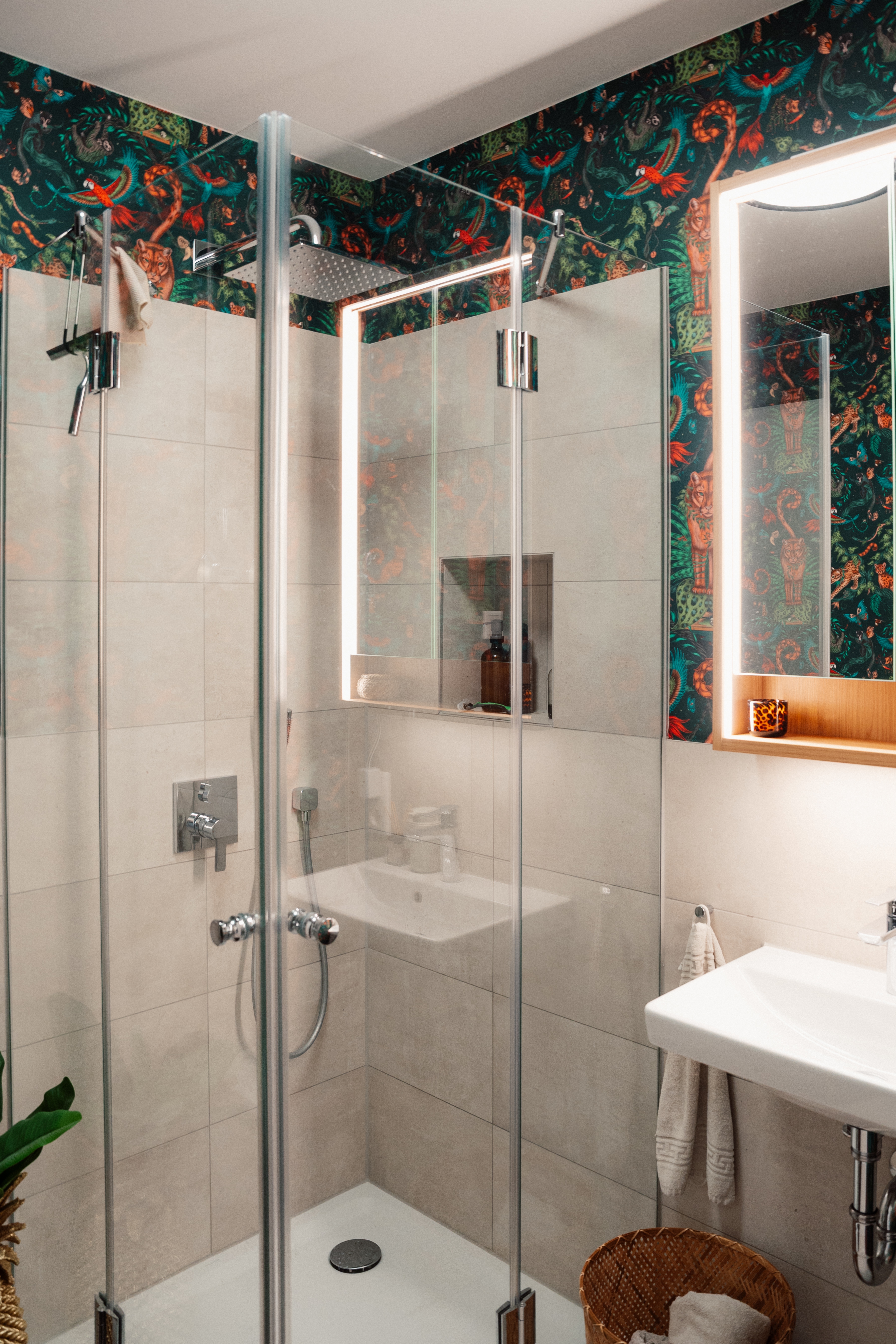 Kleines Bad mit gläserner Eckdusche, beigen Fliesen, beleuchtetem Spiegel und tropischer Tapete