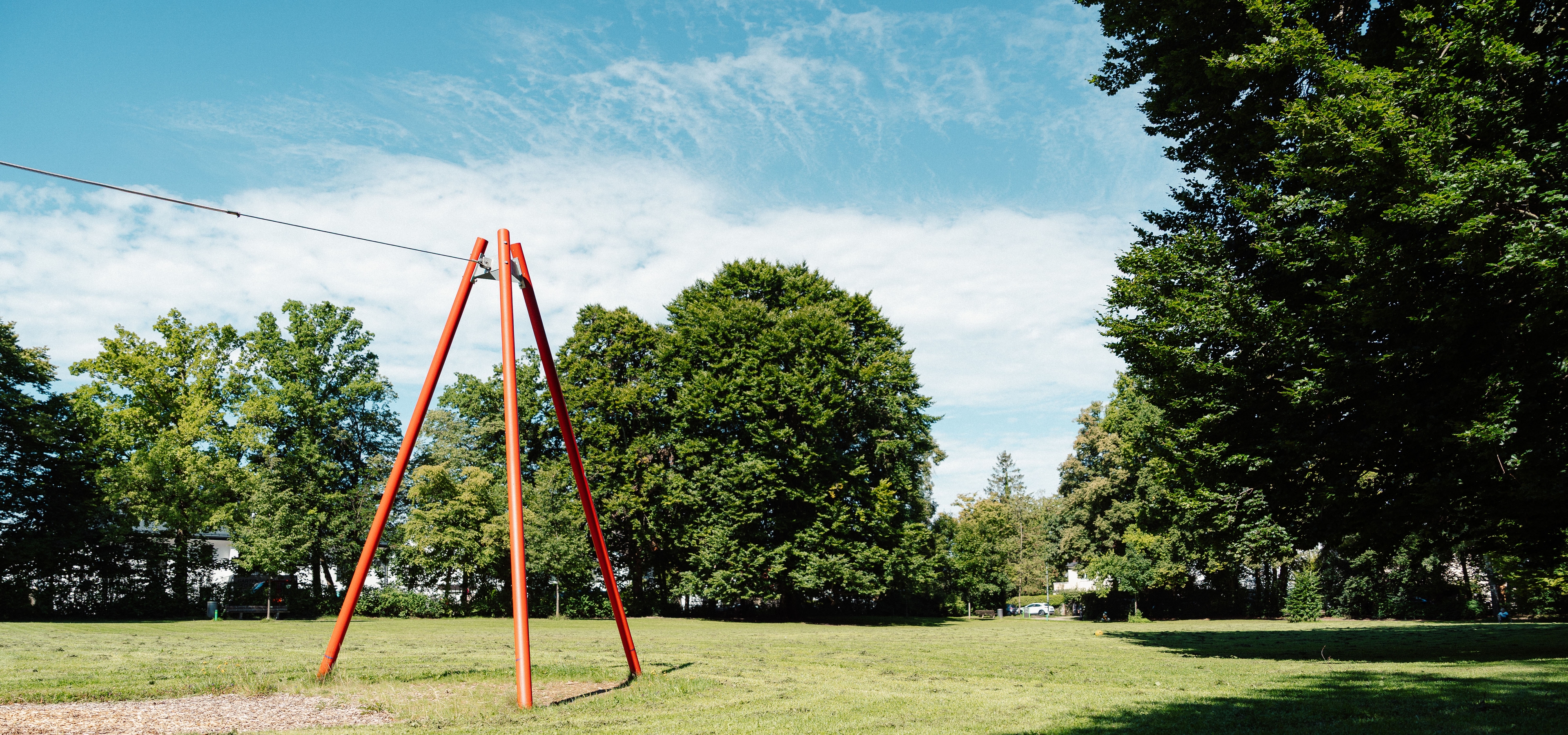 Spielplatz-Seilbahn mit roter A-Stütze auf großer Wiese im Park, umgeben von Bäumen und blauem Himmel